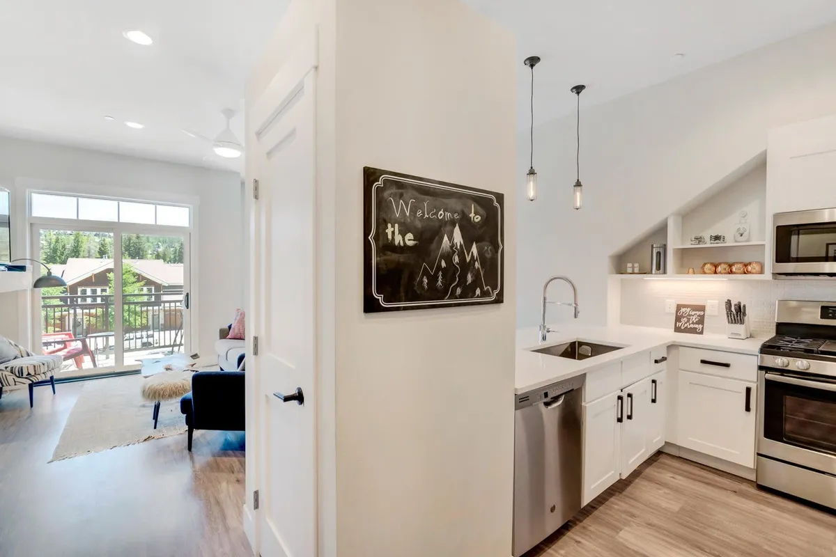 This image features a chalkboard with a welcoming message in a cozy kitchen area, visible kitchen appliances, and an open window that offers a view to the outdoors. The overall mood suggests hospitality and a sense of arrival, with morning light spilling in and hints of the landscape beyond.