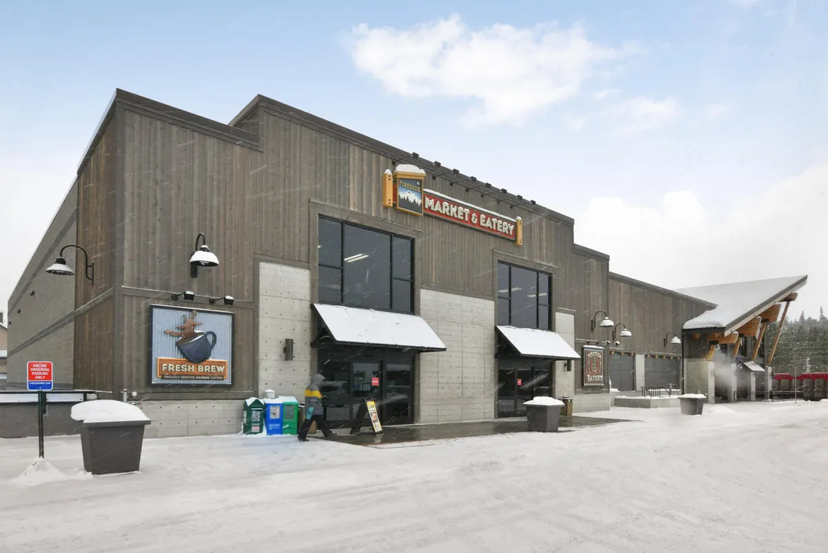 An inviting exterior view of a local market entrance, surrounded by snow-covered ground. Warm light spills from the open doors, and signs for fresh coffee and local goods hint at community gathering and off-slope experiences.
