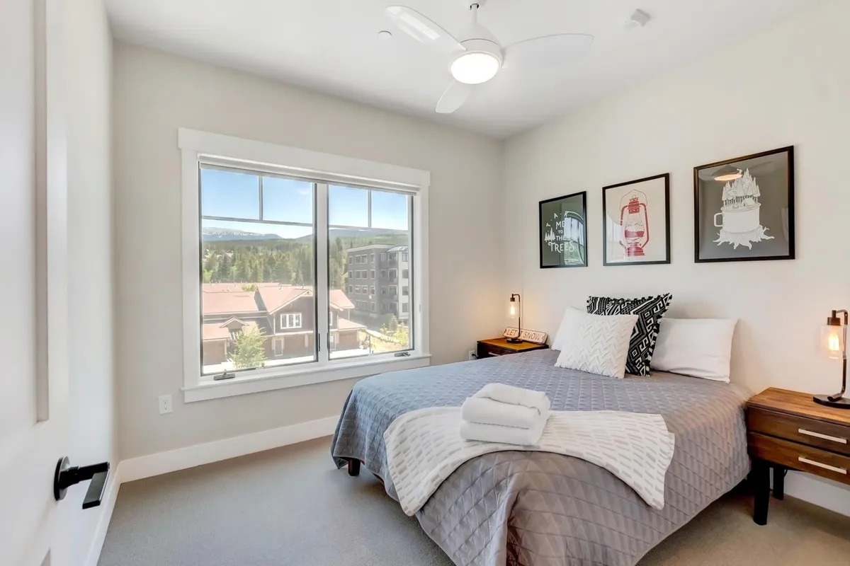 A stylish bedroom featuring a comfortable bed positioned next to a large window that offers a scenic view of the mountains outside. The decor is modern and inviting, with tasteful art pieces and a softly glowing nightstand lamp that creates a warm, welcoming ambiance.