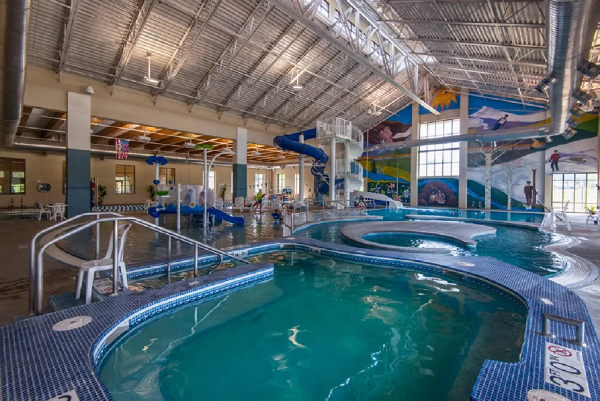 A bright, energetic indoor pool scene featuring a colorful water slide, a swimming pool filled with children playing, and a whimsical mural on the wall. The atmosphere is joyful and bustling, capturing the excitement of the Grand Park Rec Center’s family-oriented amenities.