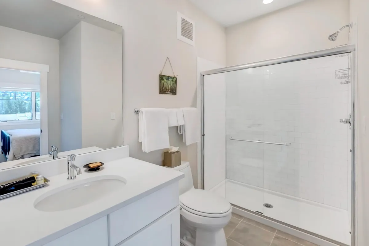A bright, modern ensuite bathroom with a vanity, large mirror, and glass shower enclosure. The design feels crisp and rejuvenating, supporting daily self-care and relaxation.