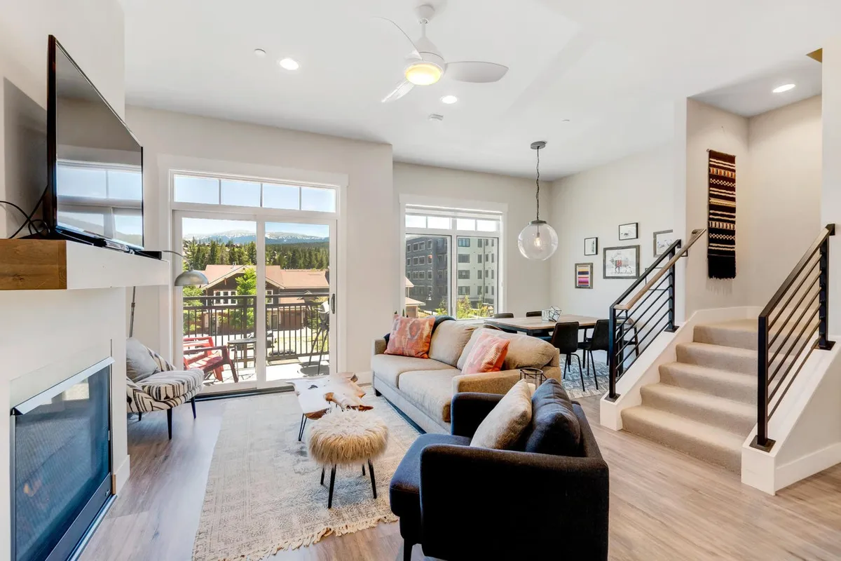 The image features a stylish, contemporary living area with a plush sofa facing a large television, a sleek dining table set for gatherings, a modern staircase, and a deck with chairs overlooking a stunning sunset. The space is bathed in natural light, blending indoor comfort with inviting outdoor views, creating an ideal setting for relaxation and connection.