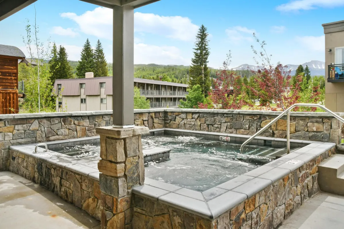 An inviting exterior view of a large hot tub nestled amid greenery and mountains, with people relaxing and enjoying the tranquil atmosphere. The spacious setting implies room for group gatherings and après-ski relaxation.