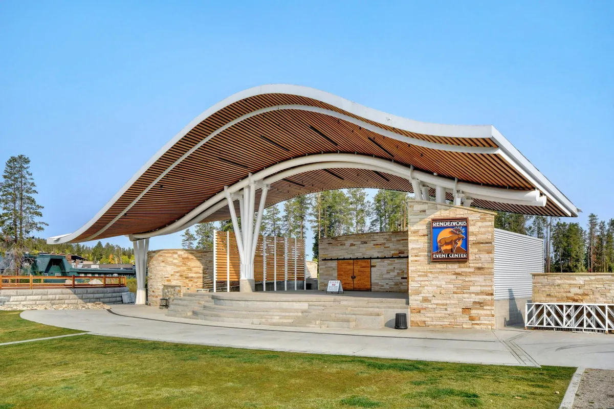 This exterior image captures the iconic curved roof structure and stone walls of Hideaway Park's amphitheater, the central outdoor venue for concerts and community events in Winter Park. The open stage and surrounding green space suggest a lively atmosphere, perfect for families attending events or enjoying the playground nearby.